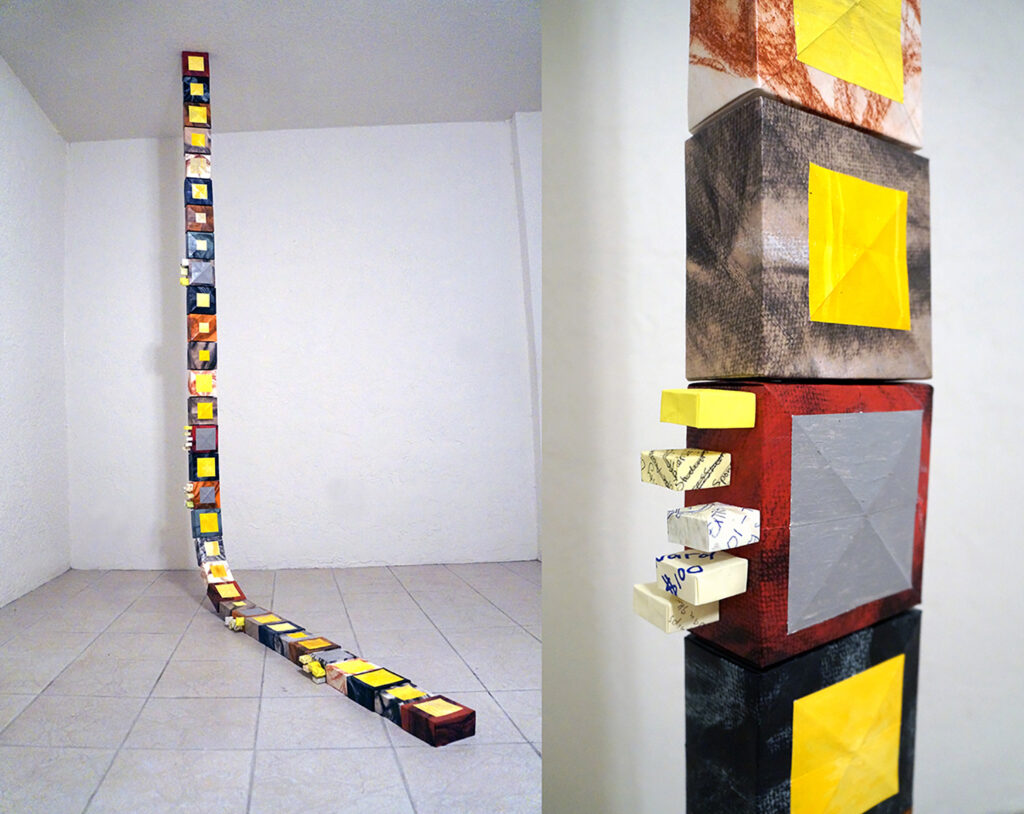 Two view of a sculpture hanging vertically from the ceiling. It is made from many small paper boxes with earthtone and gray colors. There are yellow and gray square paintings on the front.