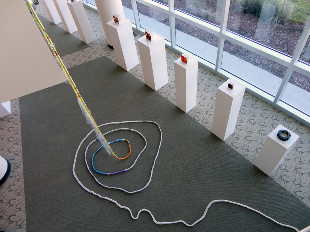Installation view inside a gallery showing a long, narrow sculpture hung from the ceiling and forming a spiral on the floor.