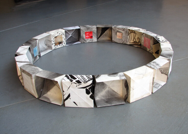 A ring of paper boxes with drawings and paintings on them, sitting on a gray floor.