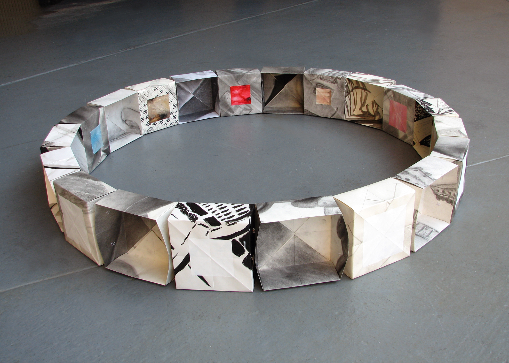 A ring of paper boxes with drawings and paintings on them, sitting on a gray floor.