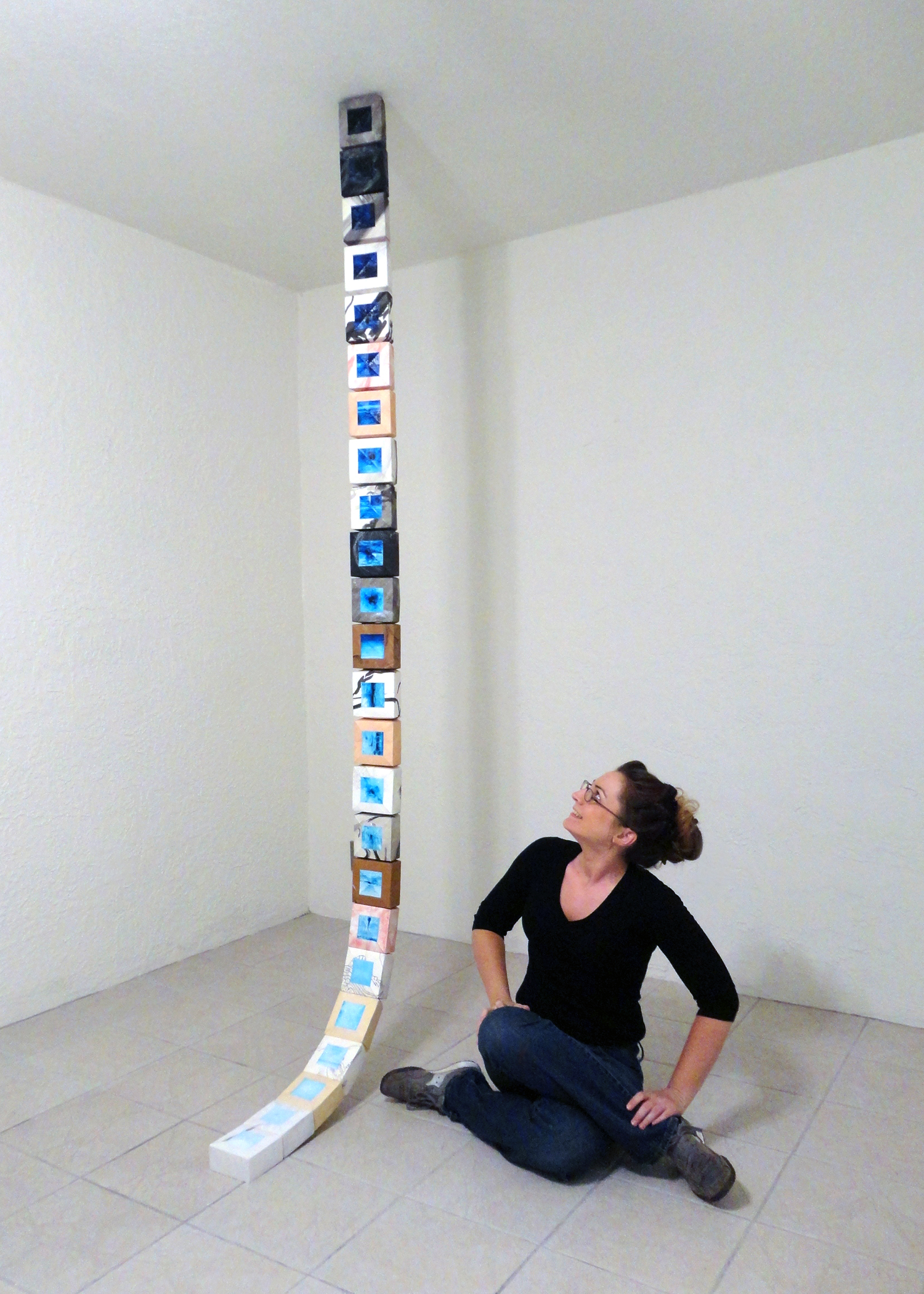 The artist Joanna White, sitting cross-legged on the floor next to her long, narrow, vertical sculpture hung from the ceiling.