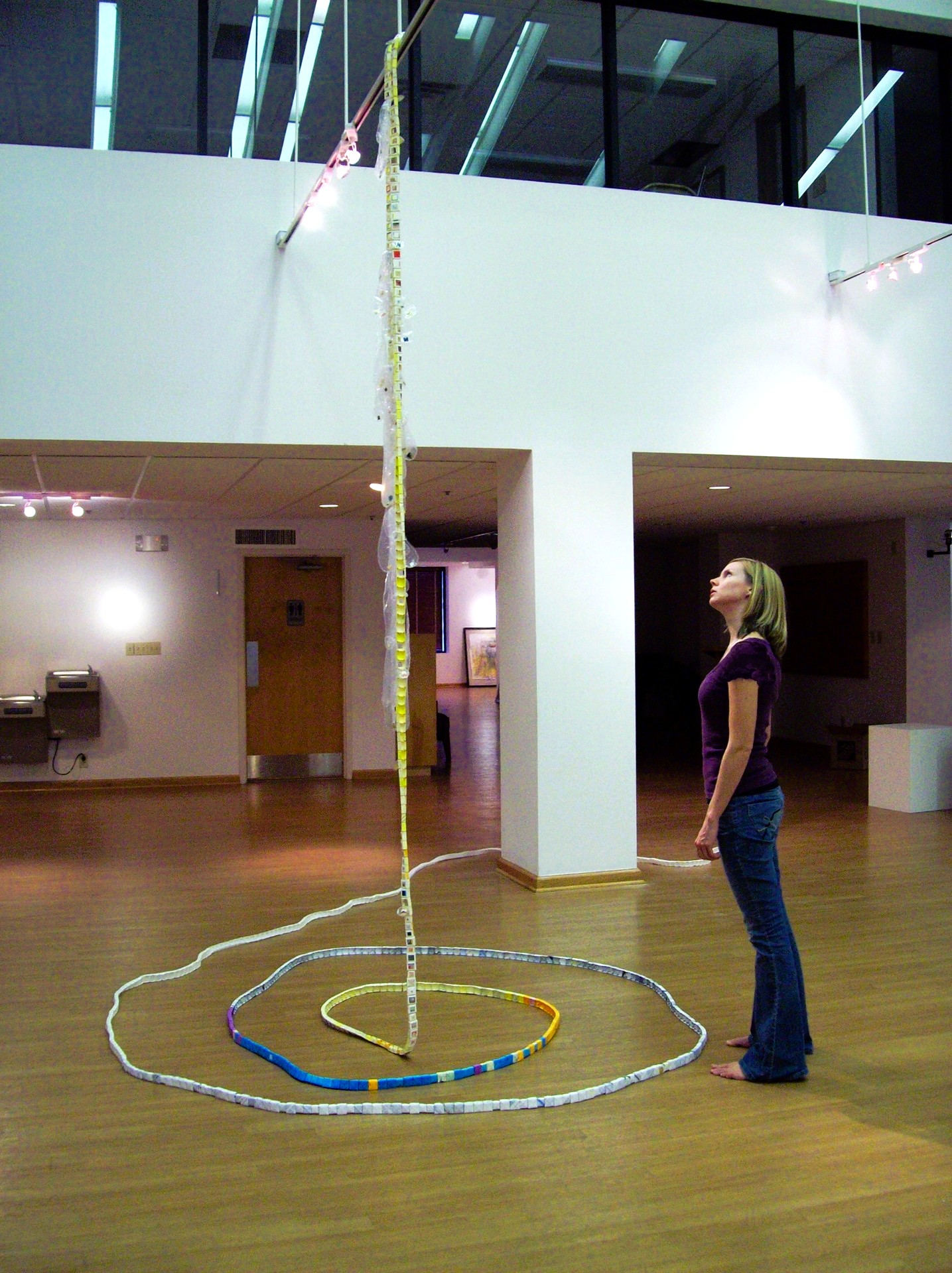 A sculpture hanging from the ceiling and forming a spiral on the floor with a gallery visitor standing next to it.