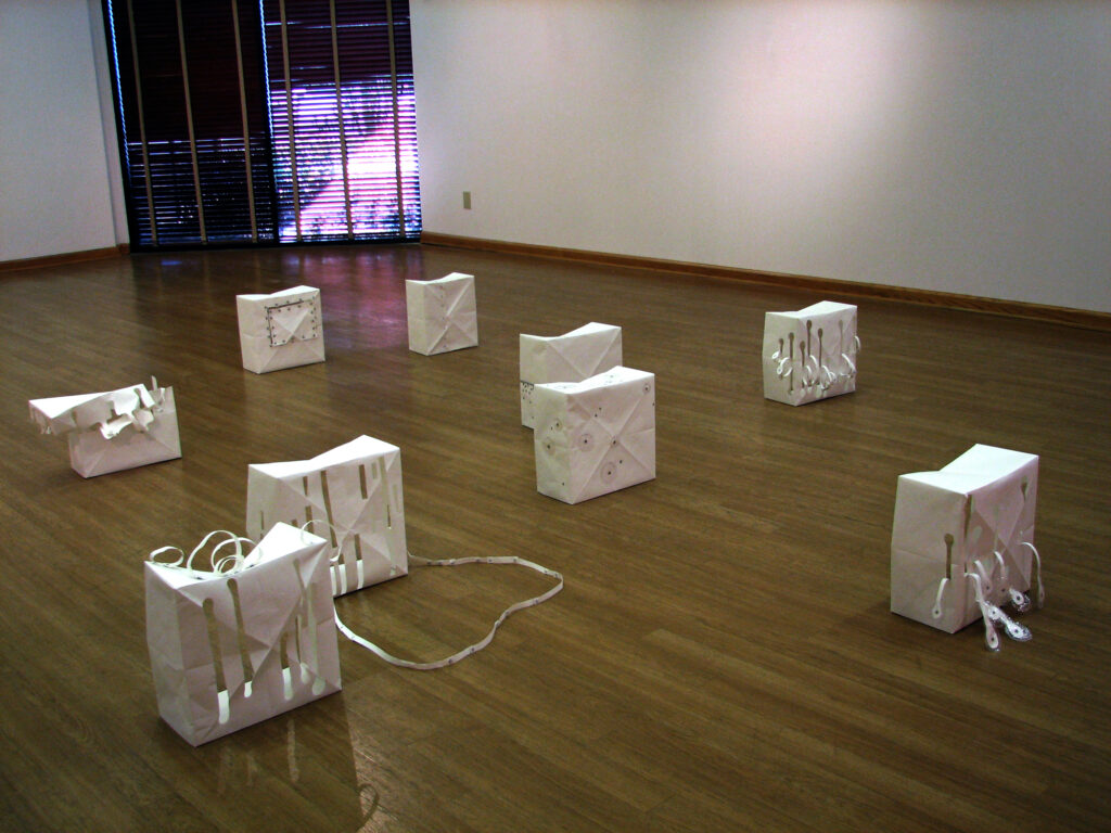 Installation art piece by artist Joanna White, featuring 9 paper boxes sitting on the floor of a gallery with various cut-outs.