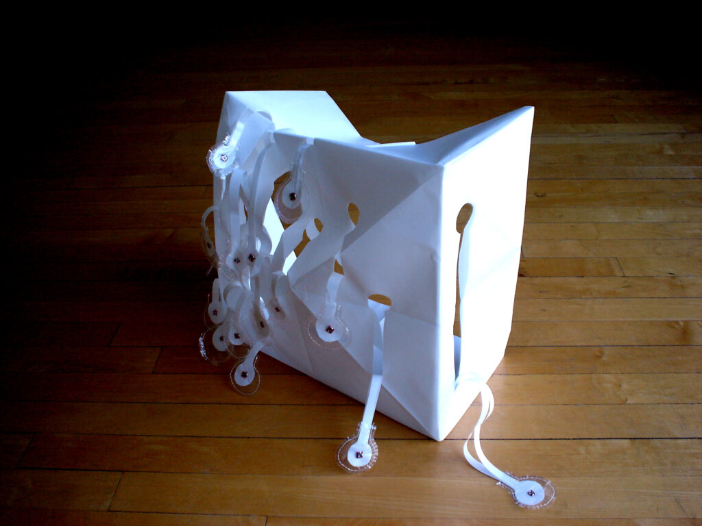 A large paper box with tabs cut out of it, stitched with metal garment snaps, sitting on a wooden floor, by artist Joanna White.
